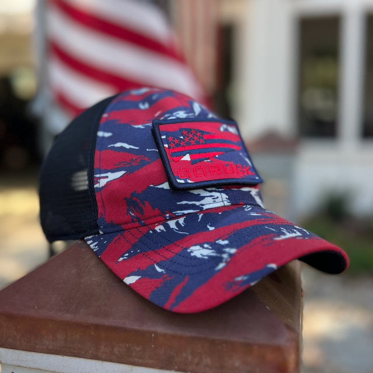 The Cadre Hat - CORDURA®, featuring a red, white, and blue camouflage pattern with a patch, is crafted from 500D CORDURA® and military-grade mesh, displayed in front of a blurred American flag and building.