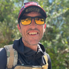 Smiling man in sunglasses and a red Cadre Hat - CORDURA® with a black jacket stands outdoors with trees in the background.