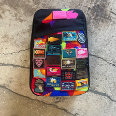 A black GR1 USA backpack made of ballistic nylon, adorned with colorful embroidered patches, rests on a cracked concrete floor.