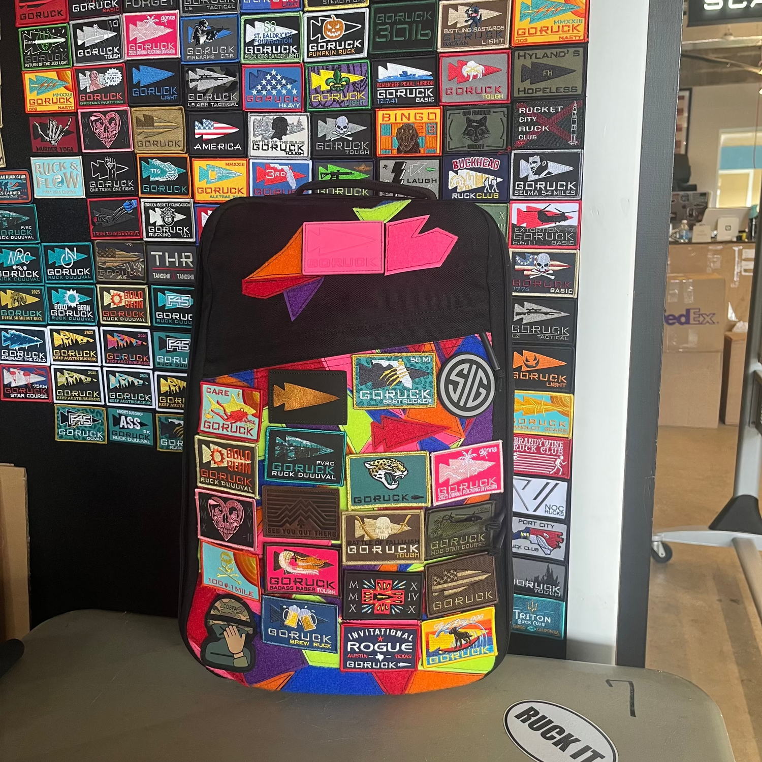 The GR1 USA backpack, made with rugged ballistic nylon and featuring a dedicated laptop compartment, is shown covered in colorful patches against a wall of similar patches.