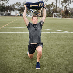 Athlete doing lunges with navy tactical rucksack overhead on green sports field with goal post