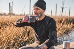 A bearded man in outdoor clothing sips from a mug while sitting in a grassy field, wearing the Men’s Commando Long Sleeve - Merino Wool shirt.