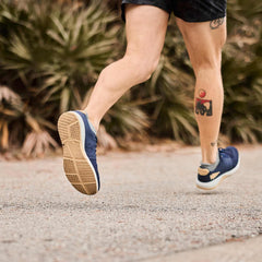 Close-up of a person running outdoors wearing blue athletic shoes and black shorts with tattoos on their legs