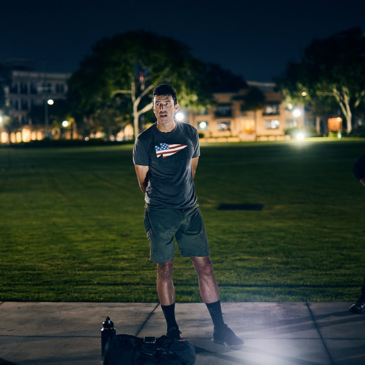 A man stands on a sidewalk at night in a grey American flag t-shirt and tough shorts, hands clasped behind his back.