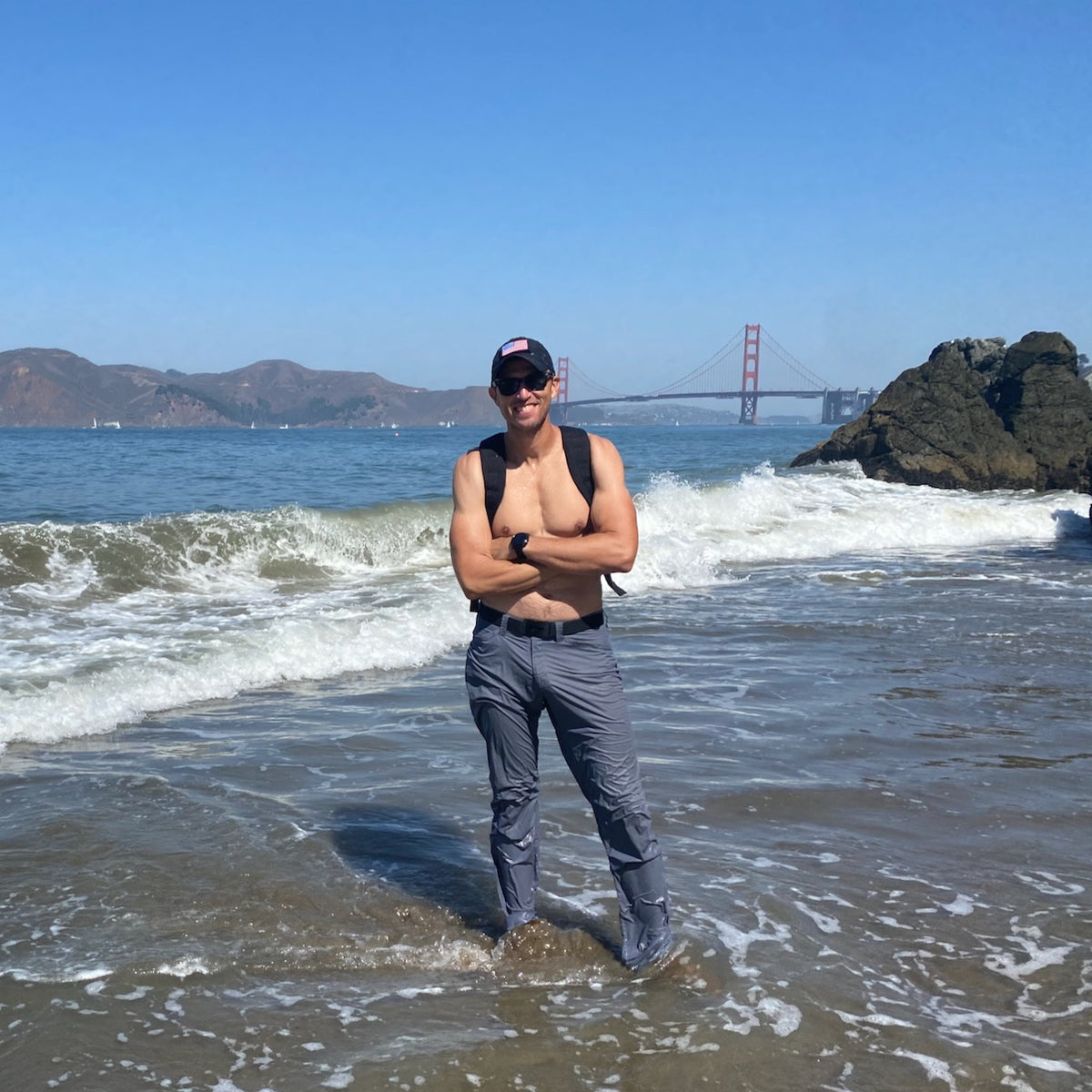 A man stands in shallow ocean water with the Golden Gate Bridge in the background on a sunny day, wearing the Spearhead Nylon Webbing Belt—an adjustable belt designed for comfort and durability.