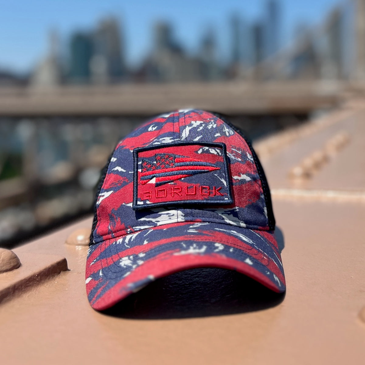 A red, blue, and white camouflage Cadre Hat - CORDURA® with an included patch, constructed from durable 500D CORDURA® fabric and military-grade mesh, is placed on a tan metal surface outdoors.