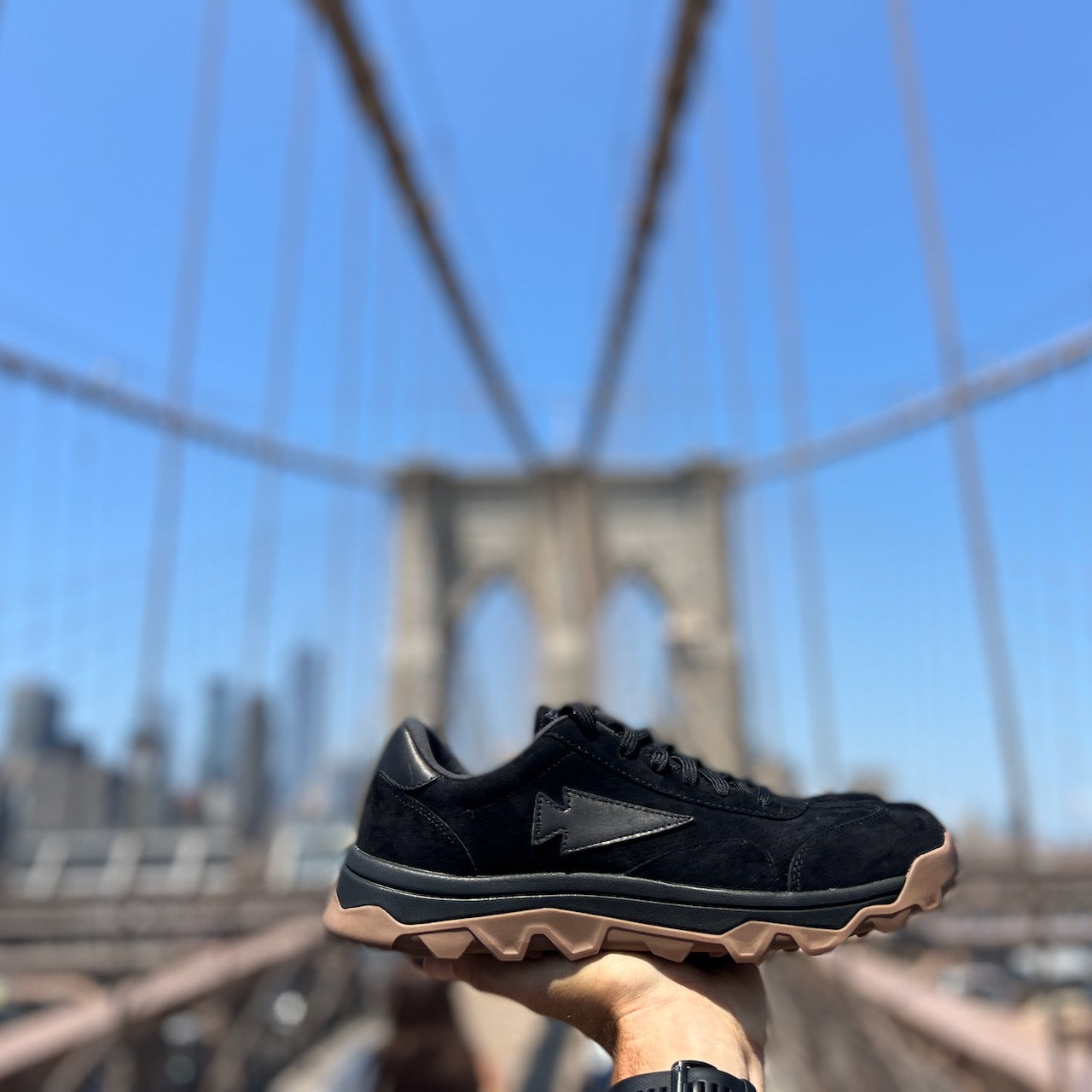 A hand holds the Mackall 45 Black + Gum sneaker in focus, with the Brooklyn Bridge blurred in the background under a blue sky.