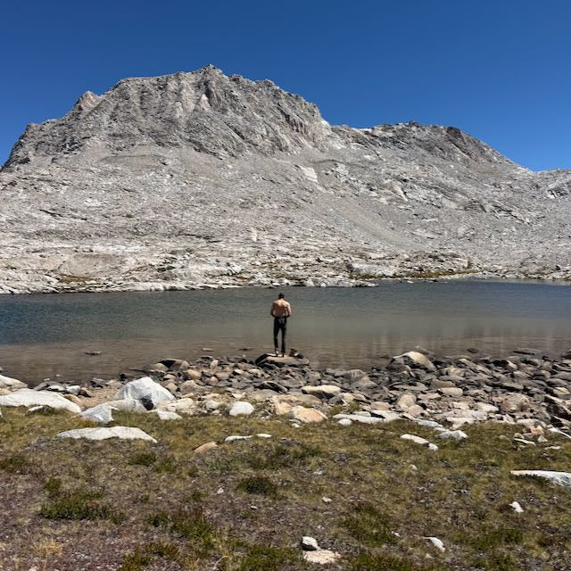 A shirtless hiker wearing fast-drying technical ToughDry pants stands on the rocky shore of an alpine lake beneath a large, barren mountain.