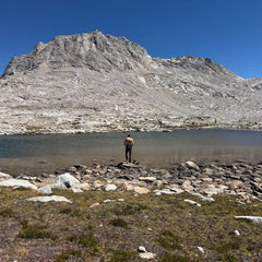 A shirtless hiker wearing fast-drying technical ToughDry pants stands on the rocky shore of an alpine lake beneath a large, barren mountain.