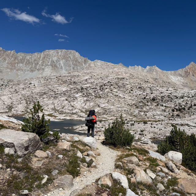 A hiker wearing ToughDry pants and a large backpack stands on a rocky trail, facing a mountain lake and rugged, treeless range under a clear blue sky.
