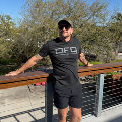 A man in sunglasses wearing a black DFQ Tee - Tri-BlendX leans on a wooden railing outdoors, with trees in the background.