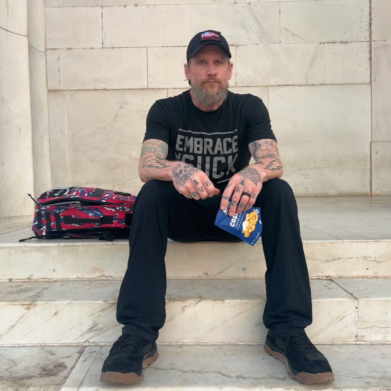 A man with tattoos sits on stone steps with a bag beside him, holding popcorn and wearing black clothes, a cap, and Mackall 45 Black + Gum sneakers.