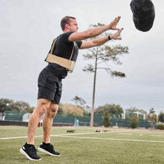 Man in rucking vest throwing a weighted sandbag outdoors on sports field with trees in background