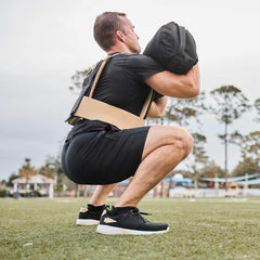 Man in black athletic wear performing a squat while holding a weighted sandbag outdoors on grass