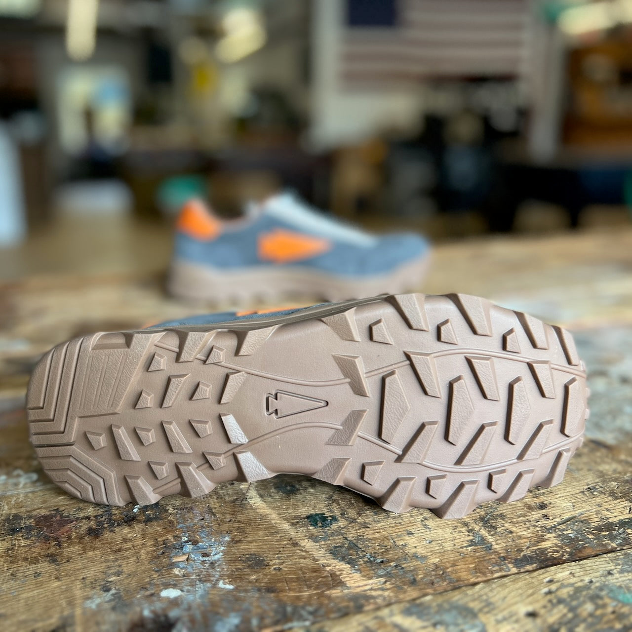 Close-up of the rugged tan sole of the Mackall 45 Fog shoe on a wooden surface, with a blurred sneaker and flag in the background.