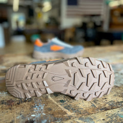 Close-up of the rugged tan sole of the Mackall 45 Fog shoe on a wooden surface, with a blurred sneaker and flag in the background.