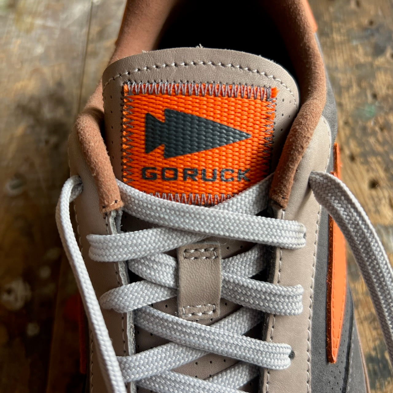 Close-up of the Mackall 45 Fog sneaker tongue featuring an orange GORUCK label and white laces, displayed on a wooden surface.