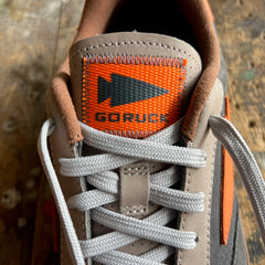 Close-up of the Mackall 45 Fog sneaker tongue featuring an orange GORUCK label and white laces, displayed on a wooden surface.