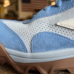 Close-up of the Mackall 45 Americana sneaker in blue and gray with a brown sole, displayed indoors against a blurred American flag backdrop.