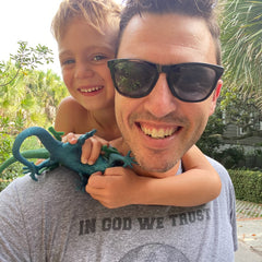 A smiling man in sunglasses with a happy child hugging him, holding the “In GOD We Trust” green toy lizard outdoors.