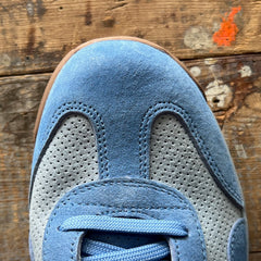 Close-up of the Mackall 45 Americana blue suede sneaker displayed on a worn wooden surface with a small orange paint spot.