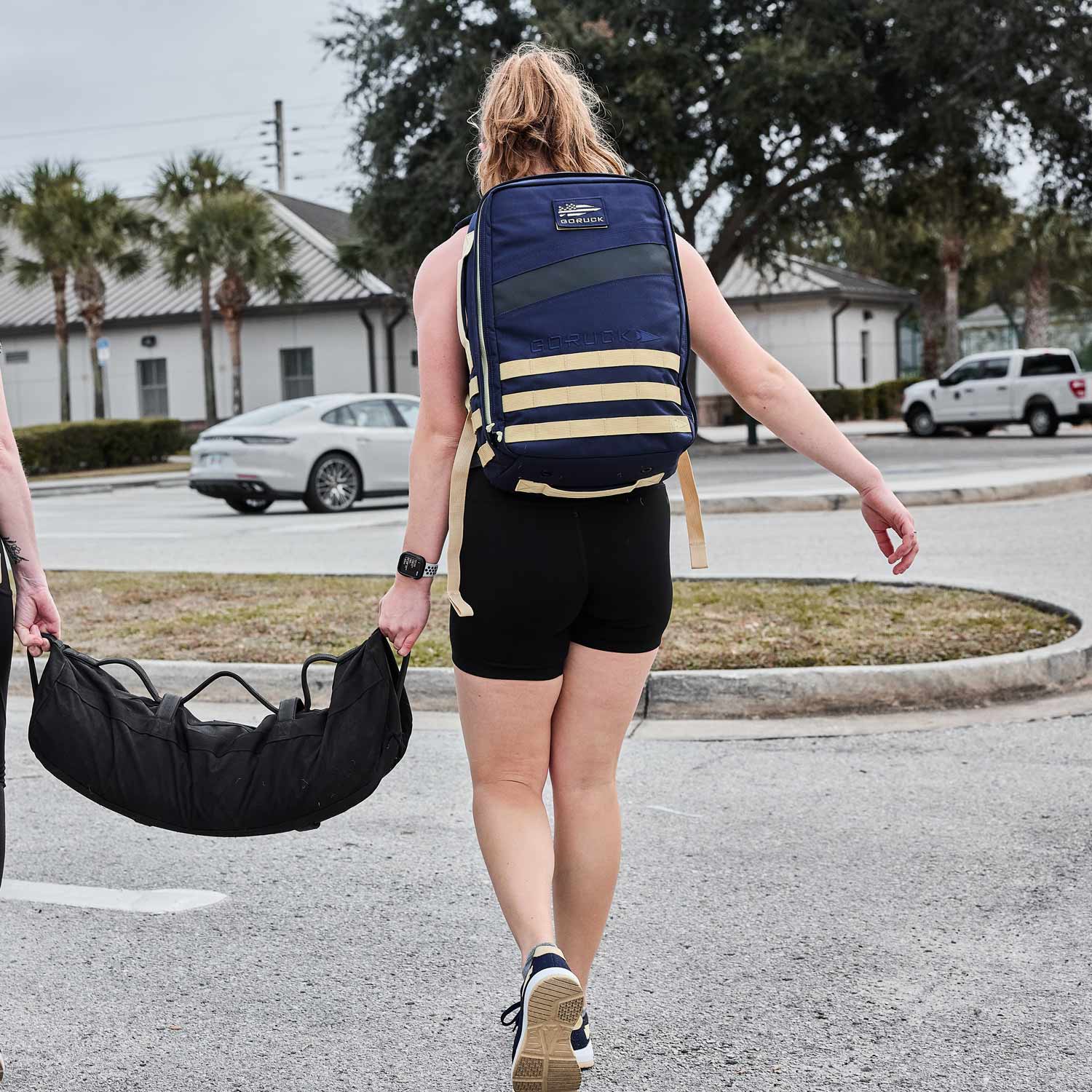 A person in athletic wear strides through the parking lot, carrying a GORUCK Rucker 4.0 loaded with ruck plates and a gym bag slung over their shoulder.