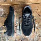Mackall 45 Black + Gum sneakers with black suede uppers and tan rugged soles displayed on a worn, paint-splattered wooden surface.
