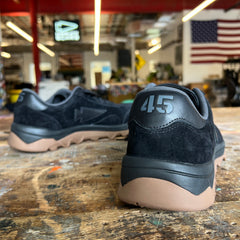 Mackall 45 Black + Gum sneakers with “415” on the heel rest on a worn table; an American flag hangs in the background.