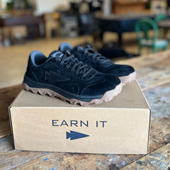 The Mackall 45 Black + Gum sneakers, featuring tan soles, displayed on a cardboard box labeled “EARN IT” with an arrow symbol.