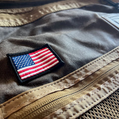 An American flag patch is sewn onto the brown waxed duck canvas of the GR1 USA - Heritage Waxed Canvas, with tan zippers and mesh details visible, evoking the rugged durability characteristic of classic heritage rucksack designs.