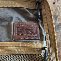 Sure! Here’s the revised sentence using your product data: A brown leather patch reading “BUILT IN THE USA HERITAGE” is featured on the tan GR1 USA - Heritage Waxed Canvas rucksack, with mesh pockets visible.