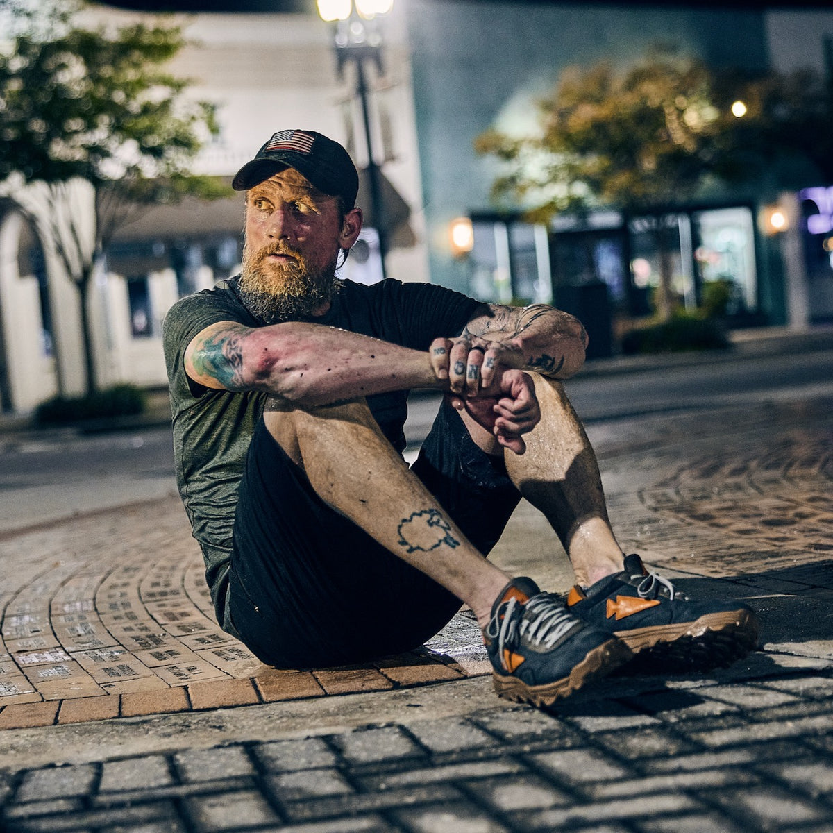 A bearded man wearing a cap and technical shorts sits on a brick-paved city street at night.