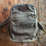 The GR1 USA - Heritage Waxed Canvas rucksack, made from brown waxed duck canvas and featuring a front pocket, rests on a rustic wooden surface.