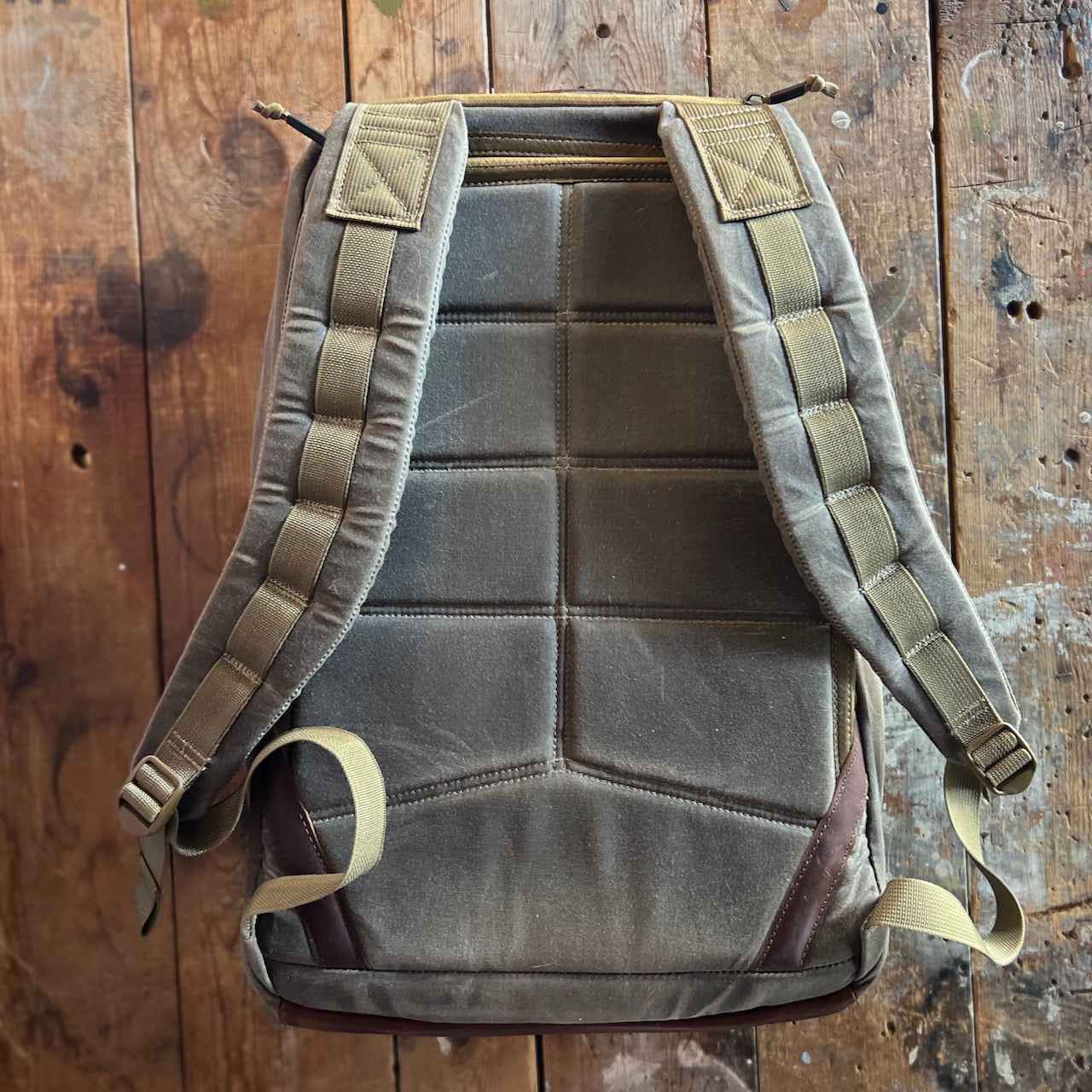 A GR1 USA - Heritage Waxed Canvas rucksack in khaki is lying flat on a rustic wooden surface, highlighting its durable waxed duck canvas construction and padded straps inspired by the iconic GR1 design.