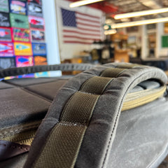 Close-up of a padded backpack strap from the GR1 USA - Heritage Waxed Canvas rucksack, with an American flag and colorful signs blurred in the background.