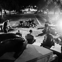 A group of men sits in a circle on a brick plaza at night during a RUCK OR DIE endurance challenge, illuminated by a bright light source.