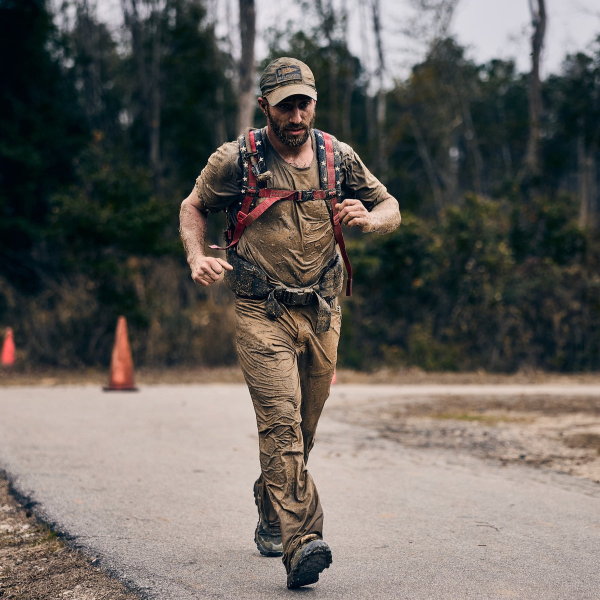 A muddy man with a beard and a baseball cap runs along a paved path wearing abrasion resistant pants and a backpack with American flag straps.