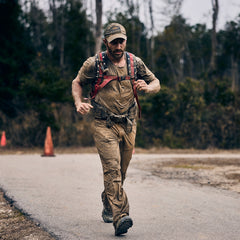 A muddy man with a beard and a baseball cap runs along a paved path wearing abrasion resistant pants and a backpack with American flag straps.