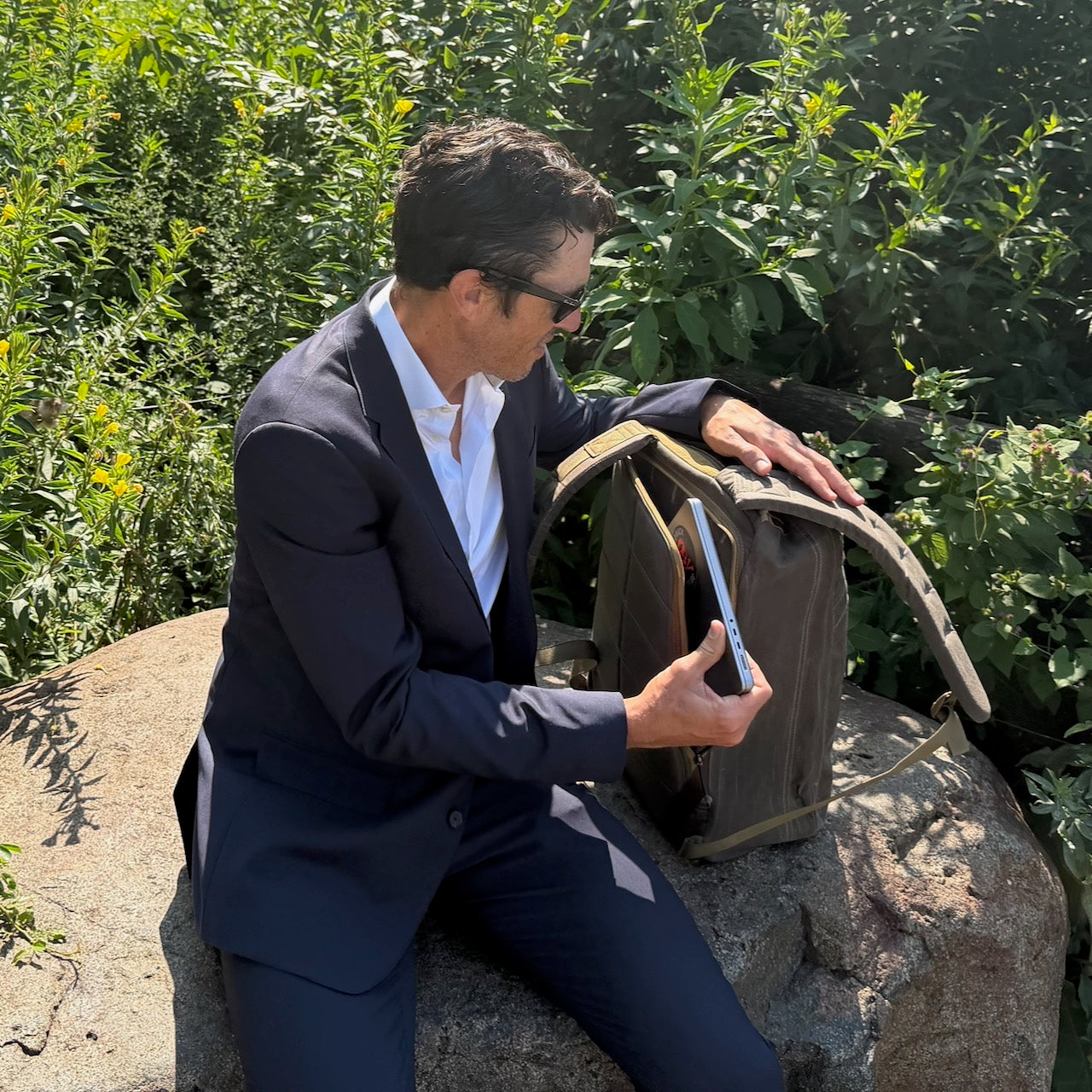 A man in a suit sits on a rock surrounded by lush greenery, opening the GR1 USA - Heritage Waxed Canvas rucksack while holding a tablet.