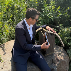 A man in a suit sits on a rock surrounded by lush greenery, opening the GR1 USA - Heritage Waxed Canvas rucksack while holding a tablet.