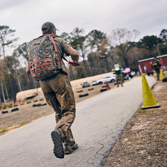 A muddy person in abrasion-resistant tactical pants and a patterned backpack runs along an outdoor path during an obstacle course race.