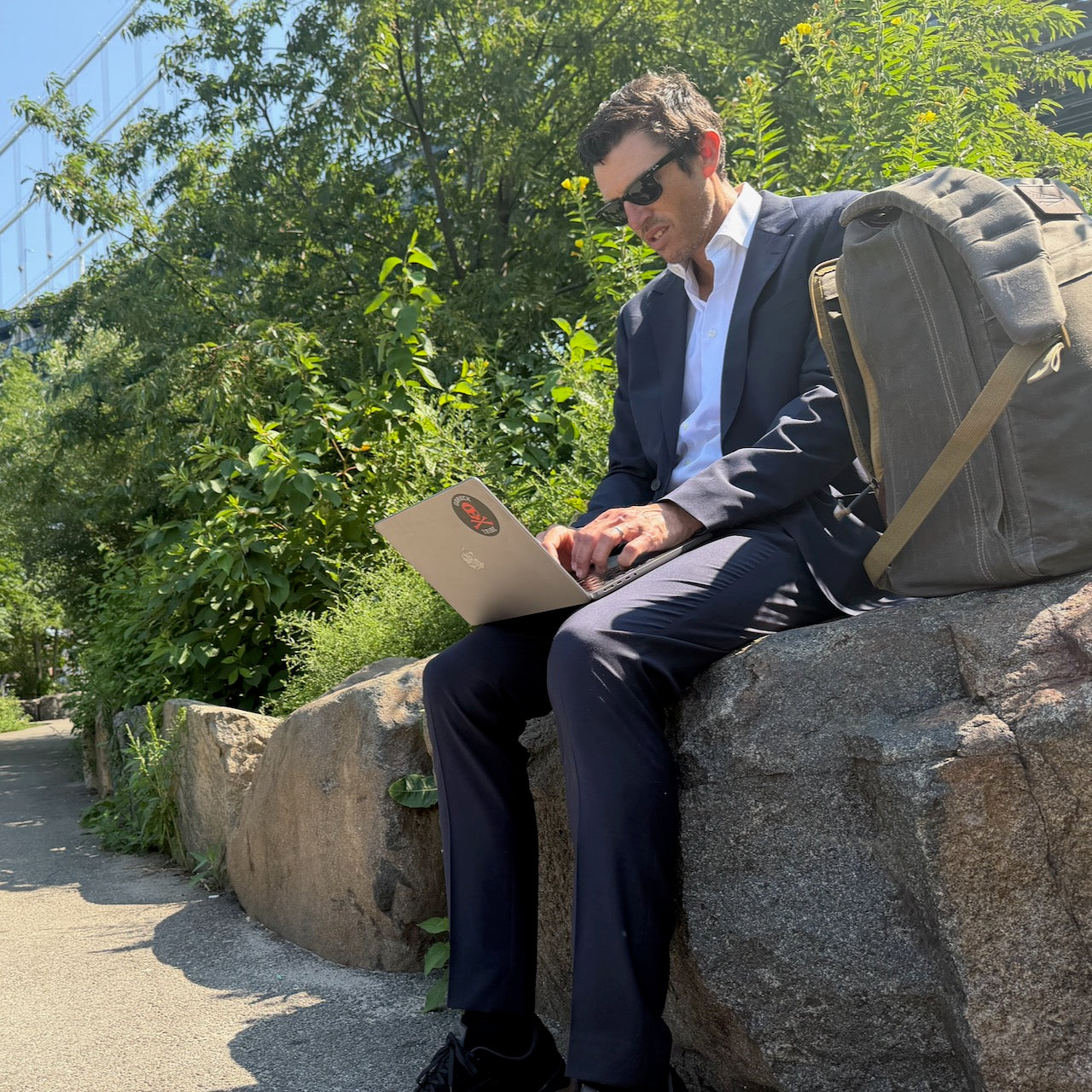 A man in a suit works on his laptop outdoors, sitting on a rock with his GR1 USA - Heritage Waxed Canvas rucksack beside him, bathed in sunlight.