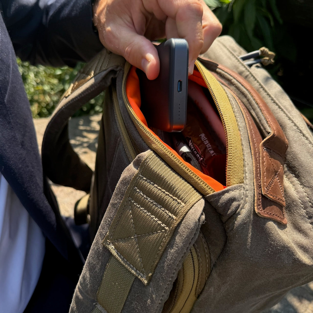 A hand places a portable charger into the front pocket of the GR1 USA - Heritage Waxed Canvas rucksack, crafted from tan and brown waxed duck canvas, outdoors.
