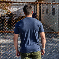 A man wearing the Slick Short Sleeve Tee - Tri-BlendX in blue and green pants stands facing a chain-link fence outdoors.