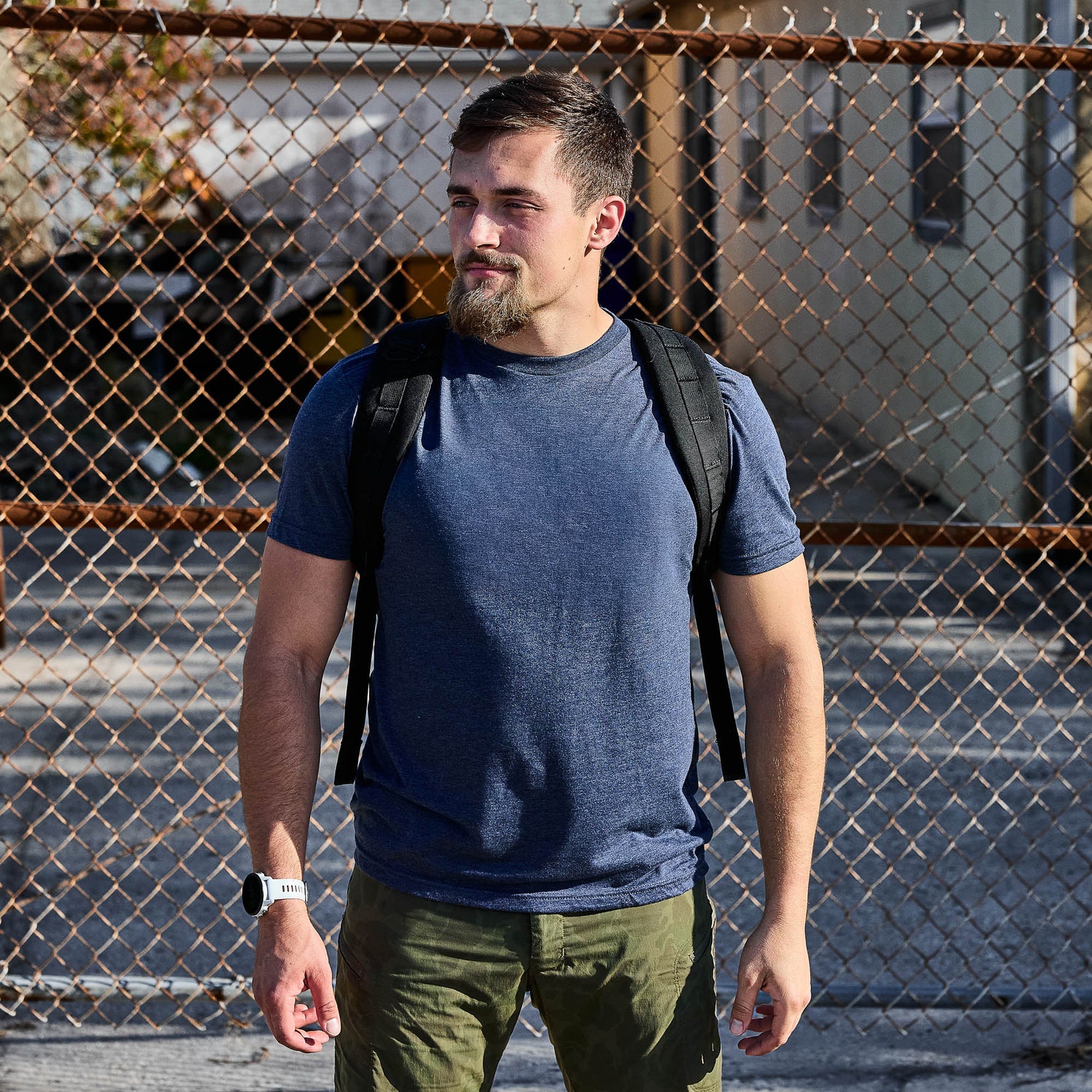 A man wearing the Slick Short Sleeve Tee - Tri-BlendX in blue and carrying a backpack stands in front of a chain-link fence on a sunny day.