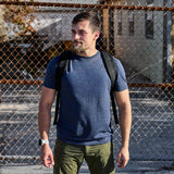 A man wearing the Slick Short Sleeve Tee - Tri-BlendX in blue and carrying a backpack stands in front of a chain-link fence on a sunny day.
