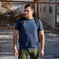 A man wearing the Slick Short Sleeve Tee - Tri-BlendX in blue and carrying a backpack stands in front of a chain-link fence on a sunny day.
