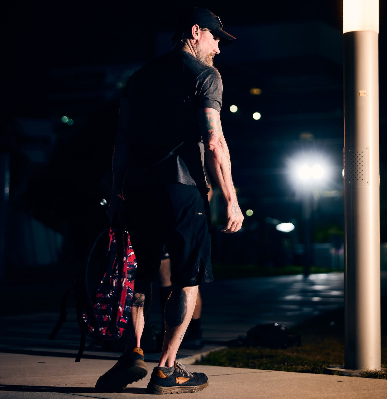 Man in shorts and cap walking at night under streetlights, carrying a duffel bag fitted with the rugged Mackall 45 Fog rucking chassis.