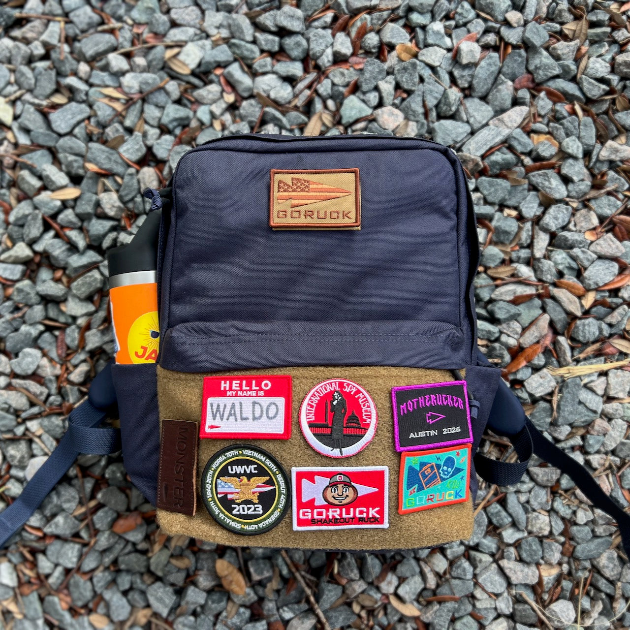 A KR1 2.0 - Kid Ruck - Add Patches, Make it Yours backpack in navy, designed for durability and customizable with colorful patches, sits on gray gravel with an orange bottle inside.
