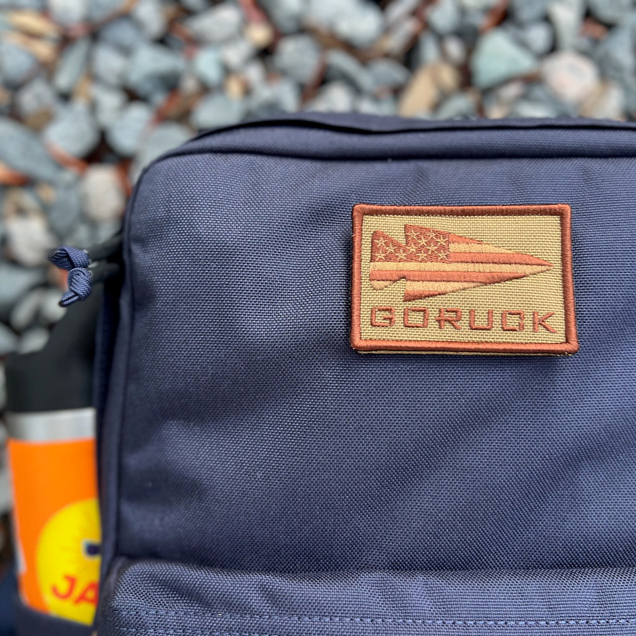 A close-up of the KR1 2.0 - Kid Ruck backpack in navy, featuring a customizable GORUCK patch and water bottle, sits on gravel—this durable kids’ backpack is ready for any adventure.